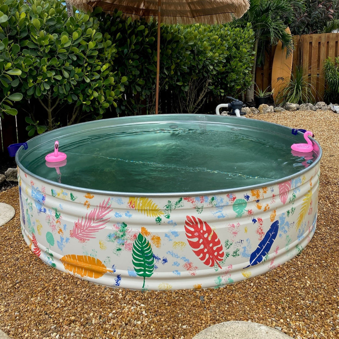 Colorful inflatable pool with tropical designs in a backyard setting with a thatched umbrella and greenery.