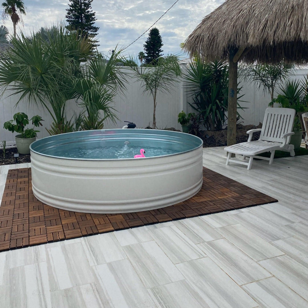 Outdoor setting with a large metal tub filled with water on a wooden deck, surrounded by plants and a thatched umbrella.
