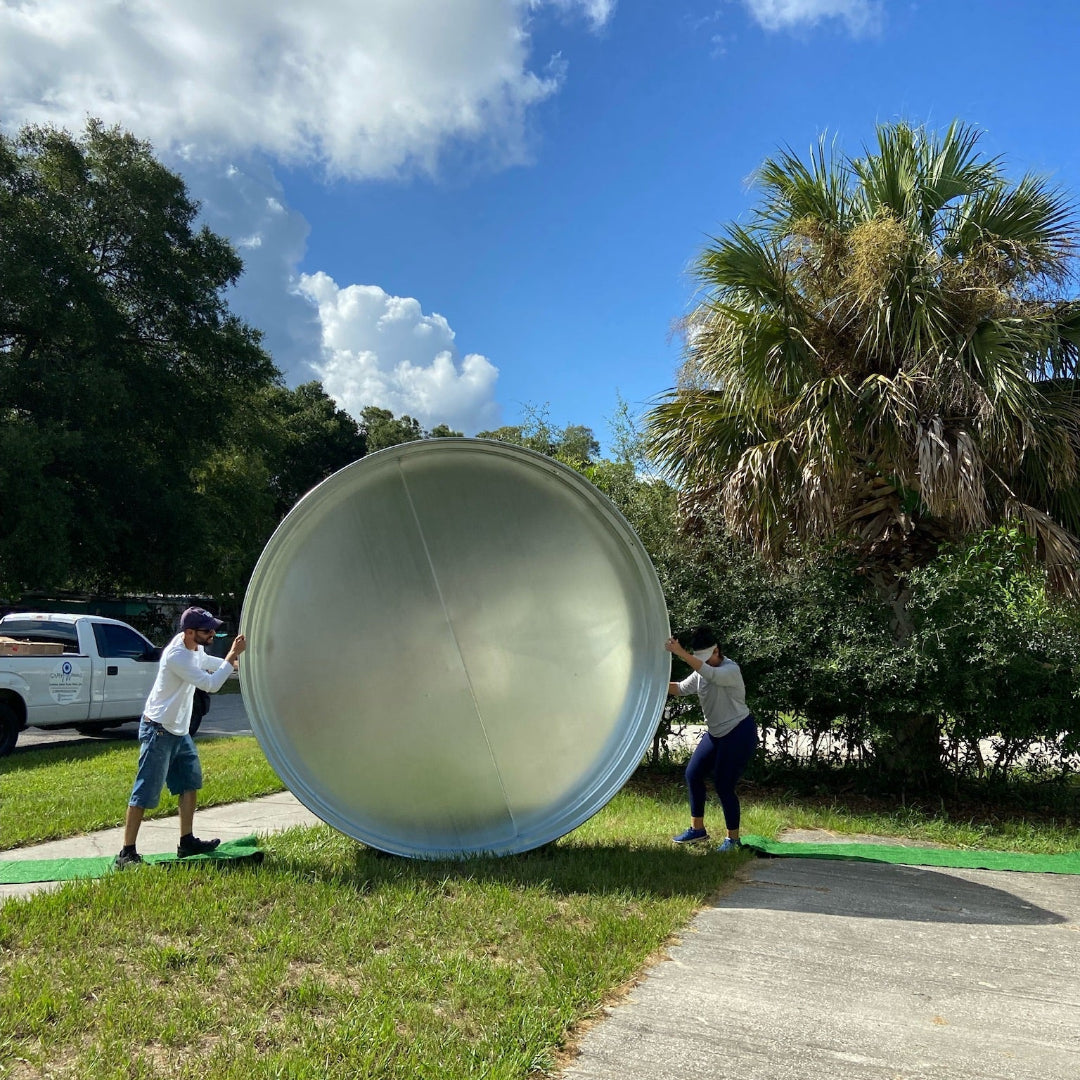 Two people moving a large cylindrical object outdoors on a sunny day with trees in the background.