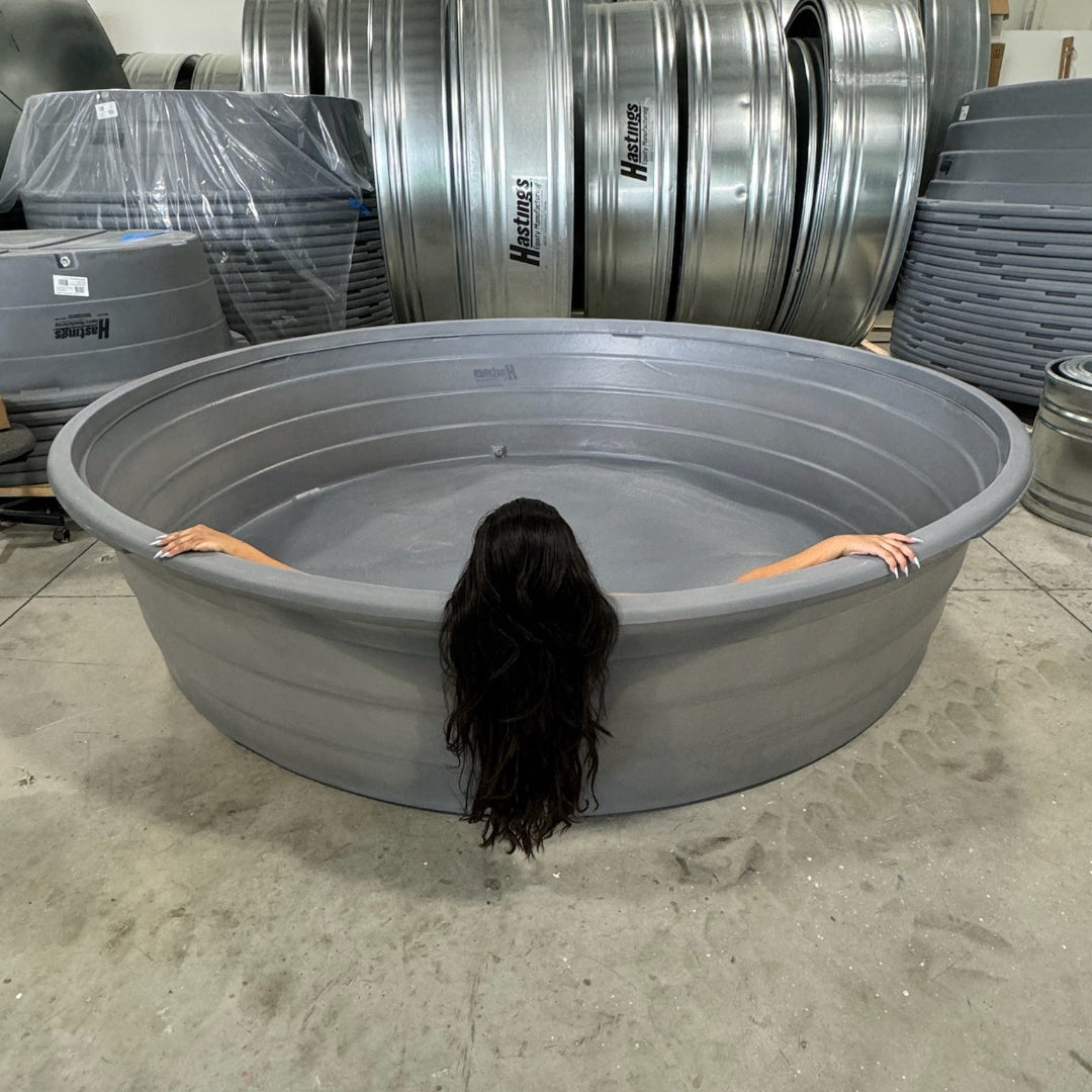 Person with long dark hair partially submerged in a large gray tub in an industrial setting with metal drums.