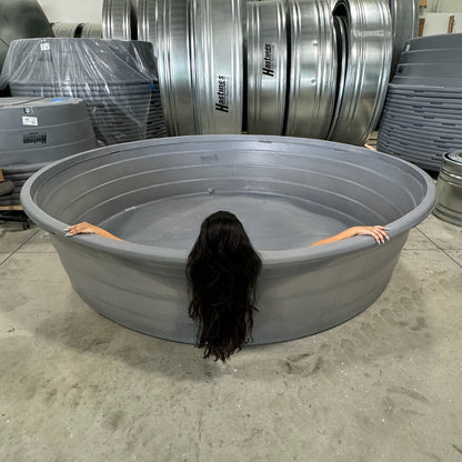 Person with long dark hair partially submerged in a large gray tub in an industrial setting with metal drums.